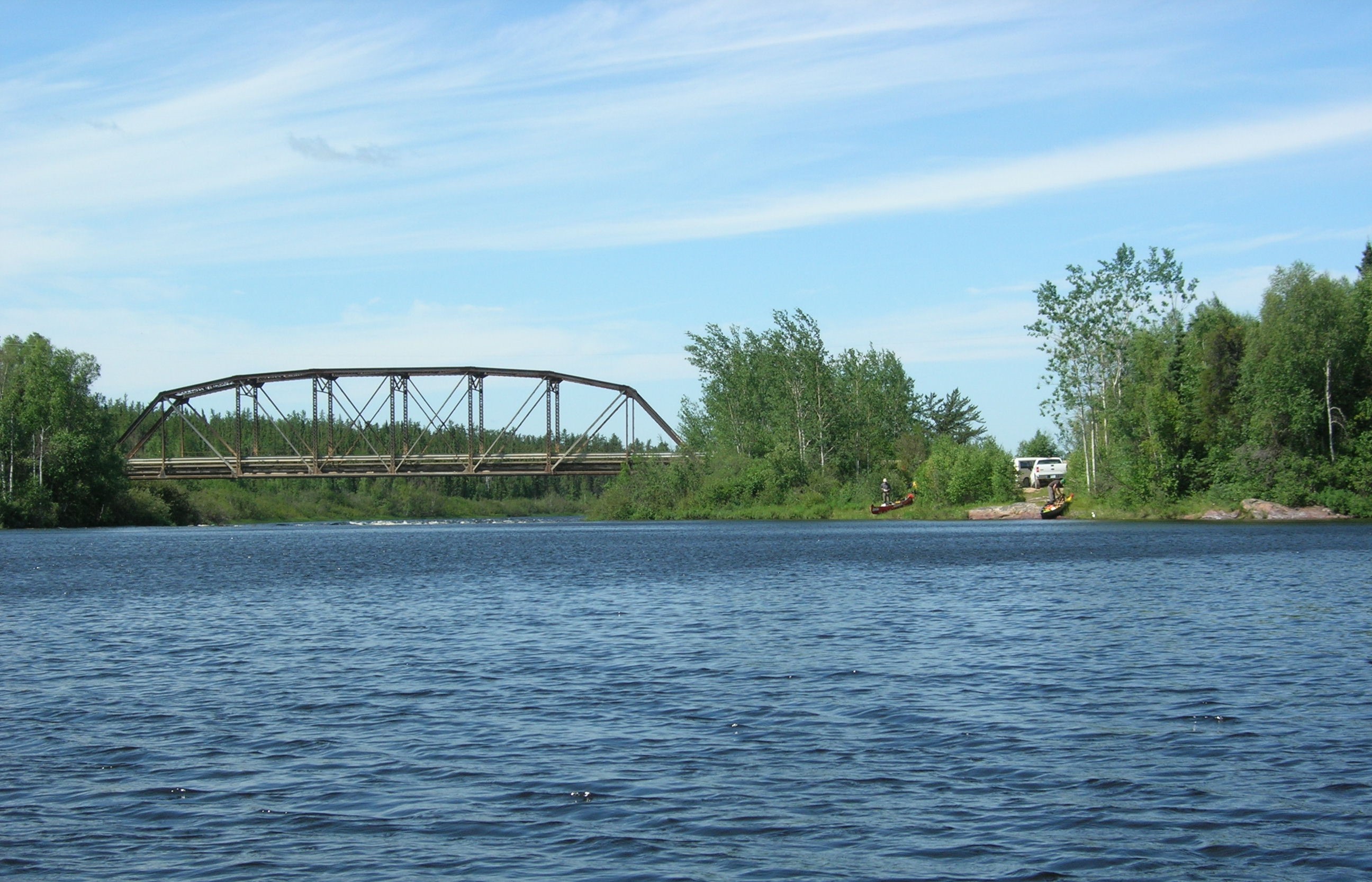 Un pont court en métal au dessus de des eaux calmes. Des rapides se dessinent au loin.