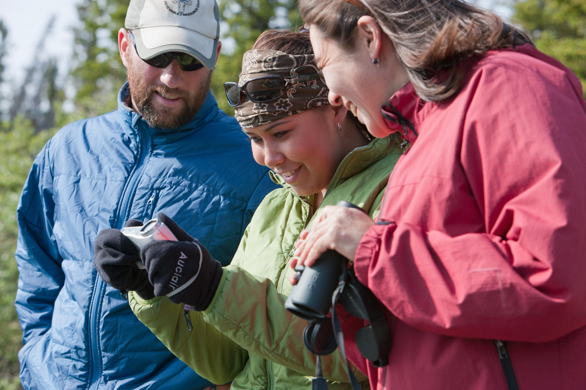 Trois adultes en tenue de randonnée regardent une photo sur un appareil photo. La personne à droite tient des jumelles.