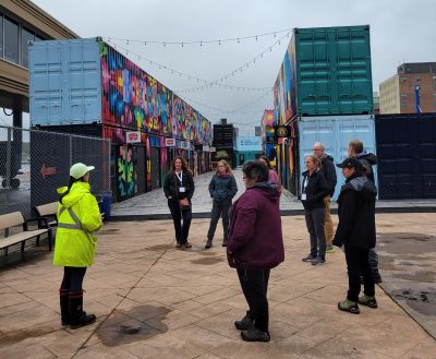 7 people standing in a circle in front of colourful containers serving as shops.