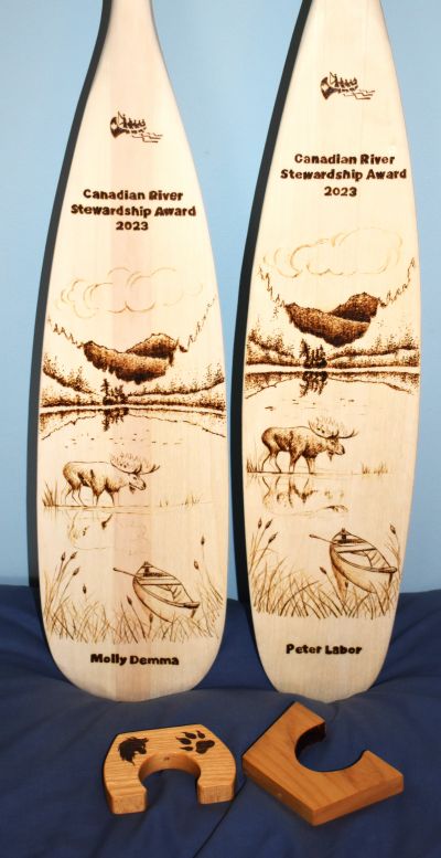 English: Two wooden paddles with a landscape, the name of the award and the name of the awardees pyrographed on them. Pyrography is the art of drawing with fire. The landscape includes a moose, forest, a canoe, and mountains. Français: Deux pagaies avec un paysage, le nom du prix et le nom des gagnants dessinés sur leur surface. Le paysage inclut un orignal, une forêt, des montagnes et un canot. Les dessins sont fait par pyrographie, soit l'art de dessiner avec le feu.