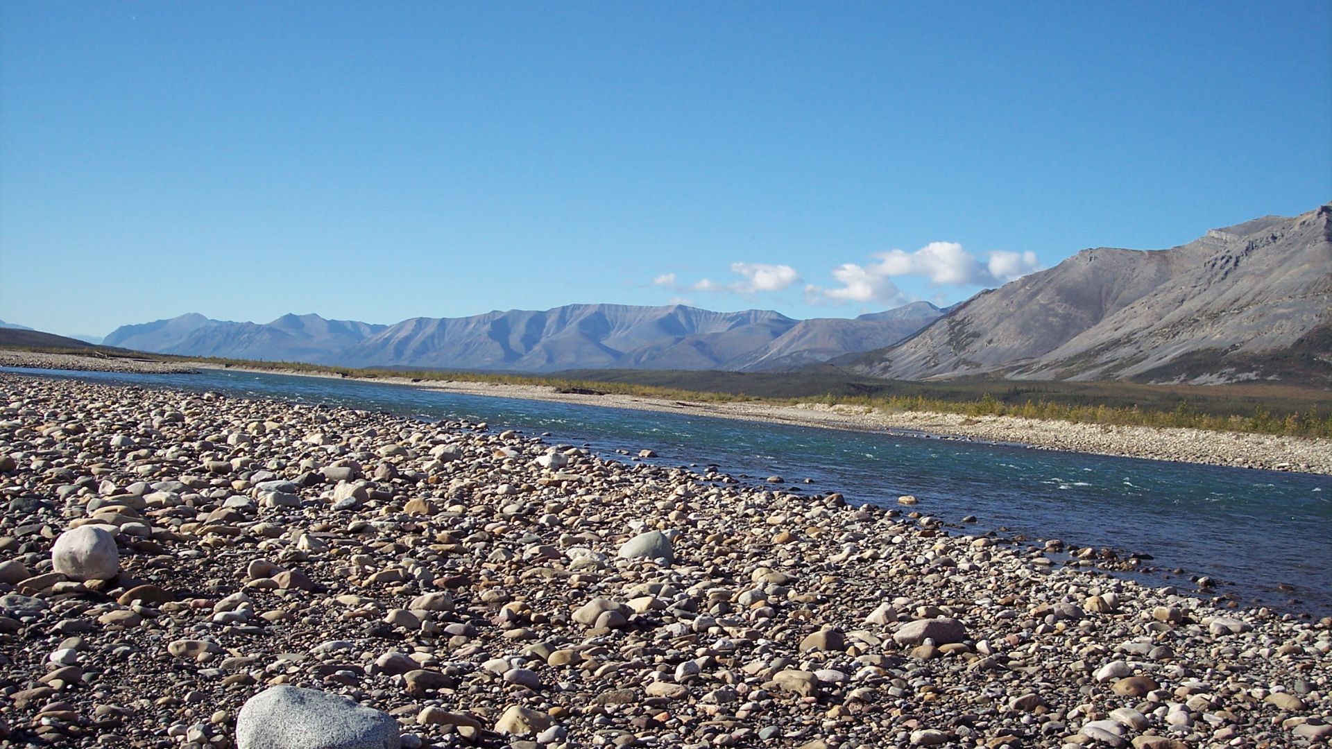 The rocky shore of the Tsiigehnjik (Arctic Red) River under a clear blue sky.