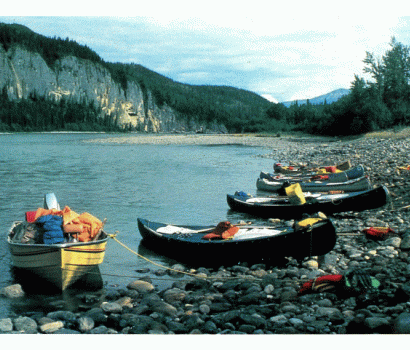 . L’image montre six canots et un bateau à moteur remplis d’équipement laissés sur le rivage rocheux de la rivière Nahanni Sud (rivière Nahʔą Dehé) pendant une partie de l’enquête sur les rivières sauvages. 